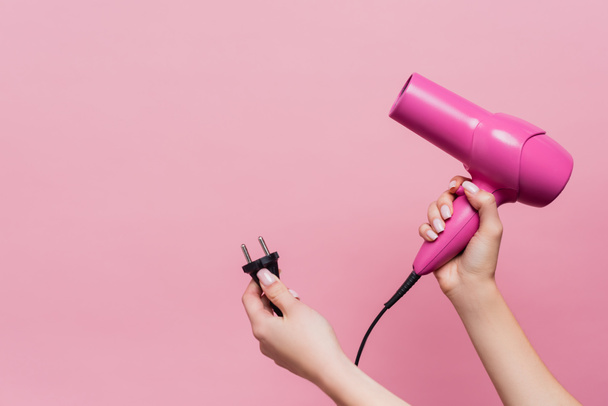 cropped view of woman holding cable with plug and hair dryer isolated on pink - Fotografie, Obrázek