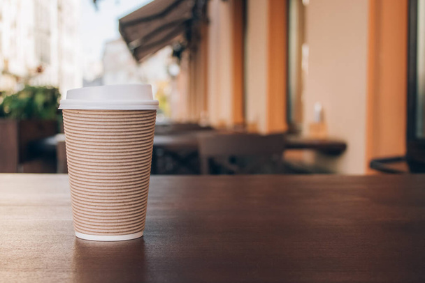 Mockup obraz papírového kelímku odnést kávu, stojící na dřevěném stole s rozmazanou kavárnou vzadu - jako pozadí s copyspace. Káva jít do recyklovatelného papírového kelímku - Fotografie, Obrázek