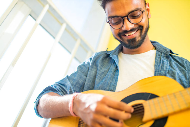 Cheerful handsome young man playing guitar and smiling in new cozy apartment - Fotografie, Obrázek