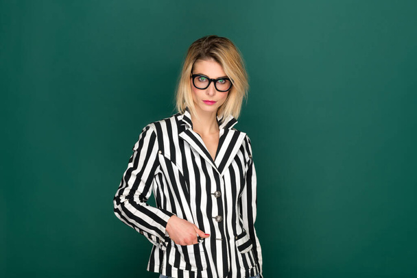 Stylish blond woman with eyeglasses in bold striped black and white jacket posing with hand in pocket over a green studio background with copyspace - Photo, image