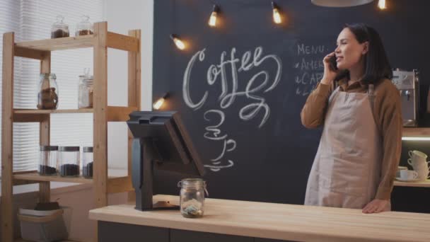Střední záběr veselý asijské prodavačka pracující v malé vnitřní kavárně s telefonem konverzace, zatímco stojí na pokladní stůl - Záběry, video