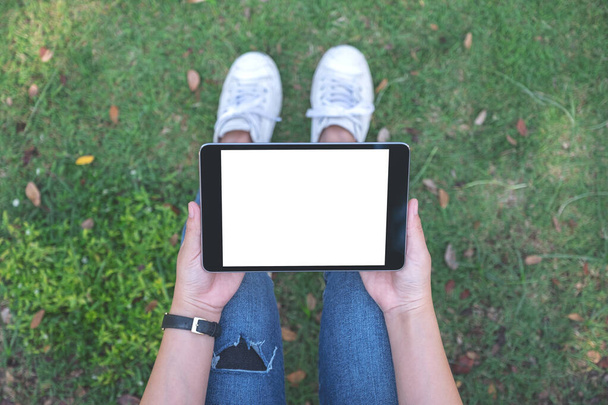 Top view mockup afbeelding van een vrouw houden en het gebruik van zwarte tablet pc met blanco witte desktop scherm horizontaal, terwijl zitten in de buitenlucht - Foto, afbeelding