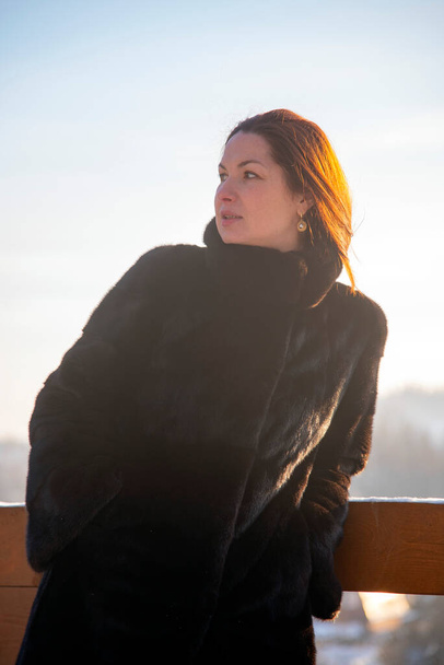 A middle-aged woman with red hair , wearing a black fur coat, against the background of a winter landscape on a sunny day. Portrait in a sunny haze. - Фото, зображення