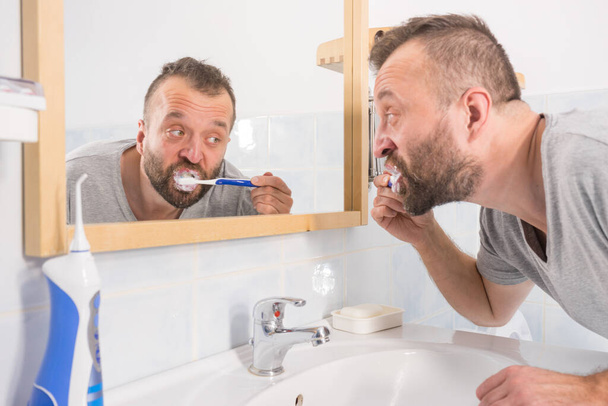 Adult man brushing his teeth looking at his bathroom mirror during morning hygiene routine. - Fotografie, Obrázek