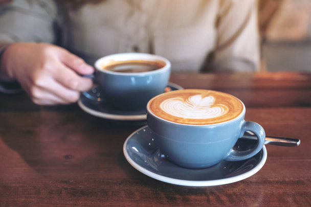 Image rapprochée d'une main tenant une tasse bleue de café noir chaud avec une autre tasse de café latte sur une table en bois dans un café - Photo, image