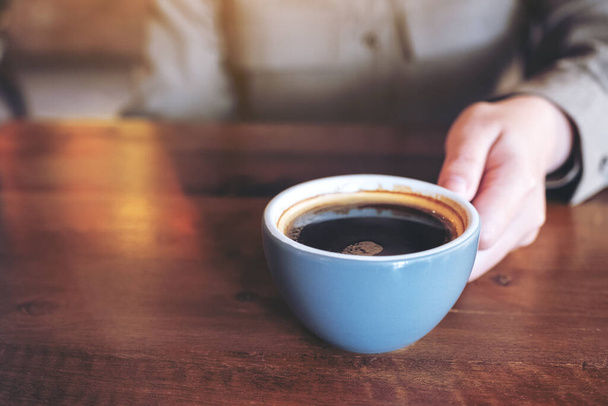 Immagine da primo piano di una mano che tiene una tazza blu di caffè nero caldo su un tavolo di legno nel caffè - Foto, immagini