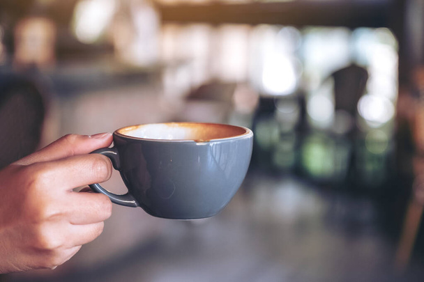 Closeup image of a hand holding a blue cup of hot coffee with blurred background in cafe - Фото, изображение