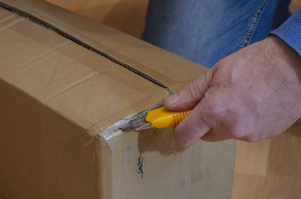 Чоловік з жовтим ножем відкриває посилку в закритій прямокутній картонній коробці на підлозі. Процес отримання онлайн-покупки. Вибірковий фокус
. - Фото, зображення