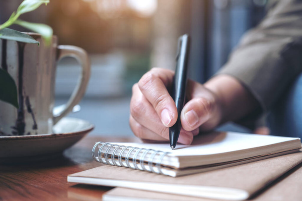 Image rapprochée de la main d'une femme écrivant sur un carnet vierge avec une tasse à café sur une table en bois - Photo, image