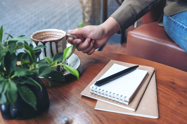 Closeup image of a hand grabbing a cup of hot coffee with notebooks on wooden table - Foto, afbeelding