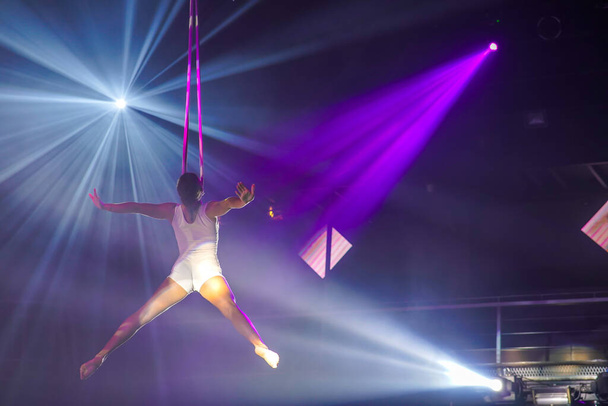 Mujer joven flexible hacer rendimiento en correas aéreas, flexible de nuevo en correas aéreas, espectáculo de circo aéreo, luz blanca púrpura. Mujer flexible gimnasta en correas - Foto, Imagen