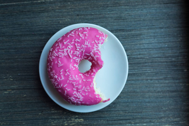 Draufsicht auf leuchtend rosa Donut auf weißem runden Teller, zweimal abgebissen. Alter hölzerner Hintergrund. Isoliertes Objekt. Traditionelle amerikanische Süßigkeiten zum Frühstück. Kopierraum. Weiße Zuckerdekoration obendrauf.  - Foto, Bild