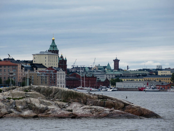 Színes épületek Helsinki belvárosában a tengertől egy szigetecskével és repülő sirályokkal az előtérben egy felhős napon. - Fotó, kép