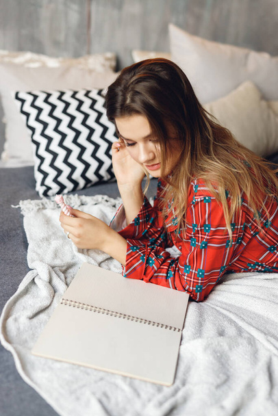 Közelről lány visel ünnepi pizsama hazugság, és írjon kívánságlistát otthon. Mosolygó nő piros hálóingben az ágyon - Fotó, kép