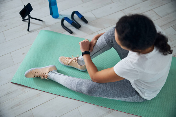 Спортсменка с фитнес-трекером сидит на коврике для упражнений - Фото, изображение
