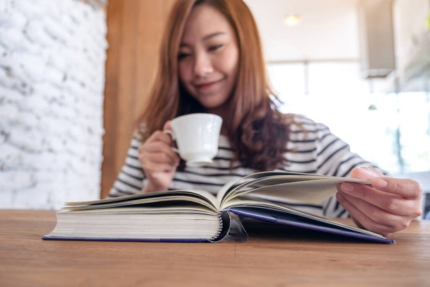 Closeup image of a beautiful asian woman holding and reading a book while drinking coffee - Фото, изображение