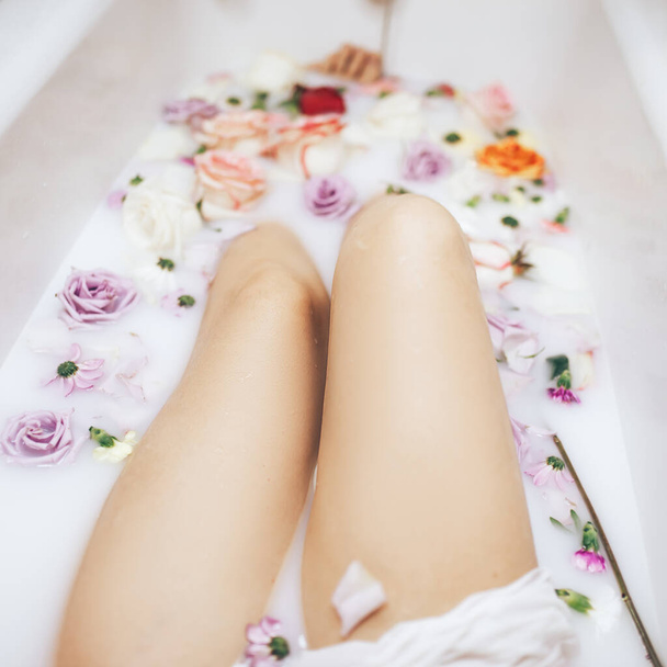 La mujer toma un baño de bienestar lleno de leche. Los brotes de rosas multicolores flotan en la superficie. Tratamientos relajantes y antienvejecimiento.. - Foto, imagen