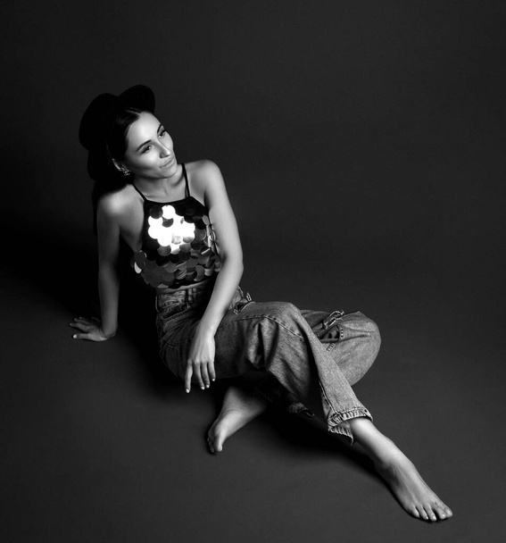 Young smiling woman in glittering top, jeans and barefoot sitting on floor and looking aside - Photo, image