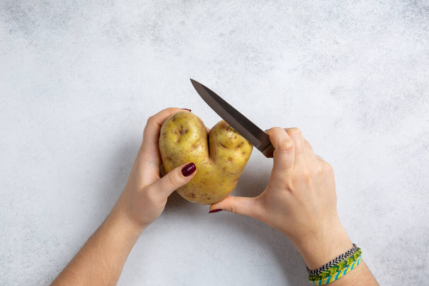 Καρδιά σχήμα πατάτας και μαχαίρι κουζίνας σε γυναικεία χέρια σε γκρι φόντο. Φαγητό μαγείρεμα, ξεφλούδισμα λαχανικών. Στο πάνω μέρος. Αντιγραφή χώρου. - Φωτογραφία, εικόνα