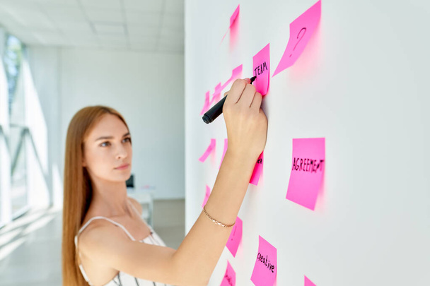 Attractive concentrated business woman using sticky notes to write and share ideas in creative office - Φωτογραφία, εικόνα