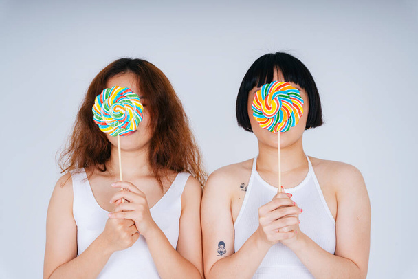 Portrait of two young asian women wearing white vest holding rainbow candy lollipop isolate on white background. - 写真・画像