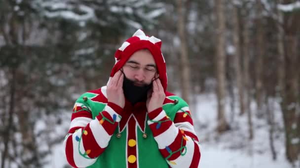 portret młodego mężczyzny w świątecznym swetrze, zakłada czarną maskę i patrzy w kamerę, w środku lasu. - Materiał filmowy, wideo