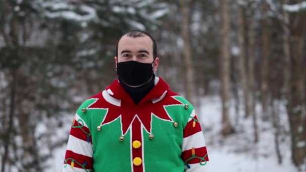 portret van een jonge man in een kersttrui, met een masker op, kijkt rond, midden in het bos. - Video