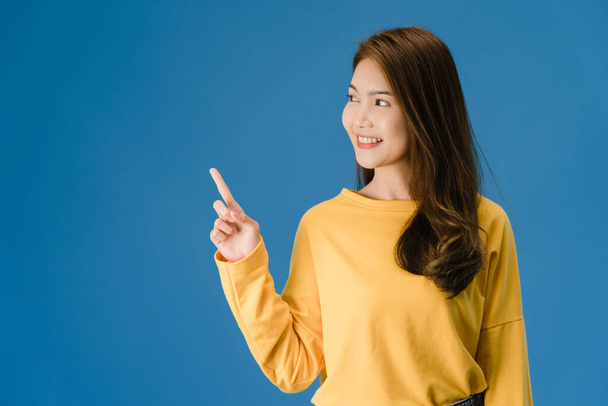 Portrait of young Asian lady smiling with cheerful expression, shows something amazing at blank space in casual clothing and standing isolated over blue background. Facial expression concept. - Photo, Image