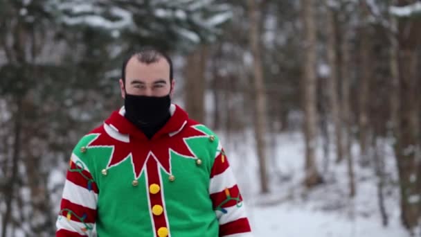 πορτρέτο ενός νεαρού άνδρα σε ένα χριστουγεννιάτικο πουλόβερ, εκτός εστίασης έρχεται στο προσκήνιο, βγάζει τη μαύρη μάσκα χαμογελώντας στην κάμερα και δείχνει την τάξη στη μέση του δάσους. - Πλάνα, βίντεο