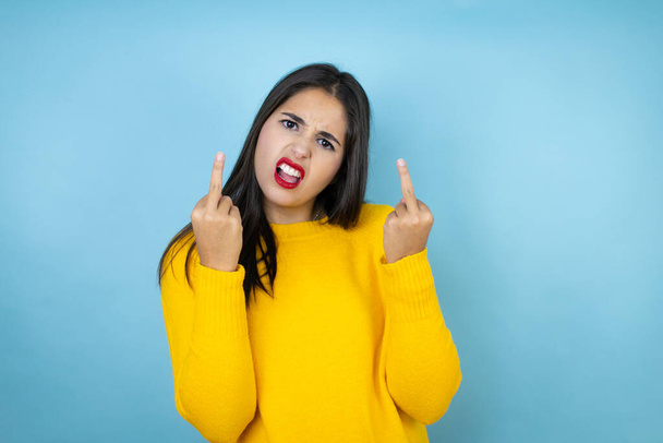 Young beautiful woman wearing yellow sweater over isolated blue background showing middle finger doing fuck you bad expression, provocation and rude attitude. screaming excited - Фото, зображення