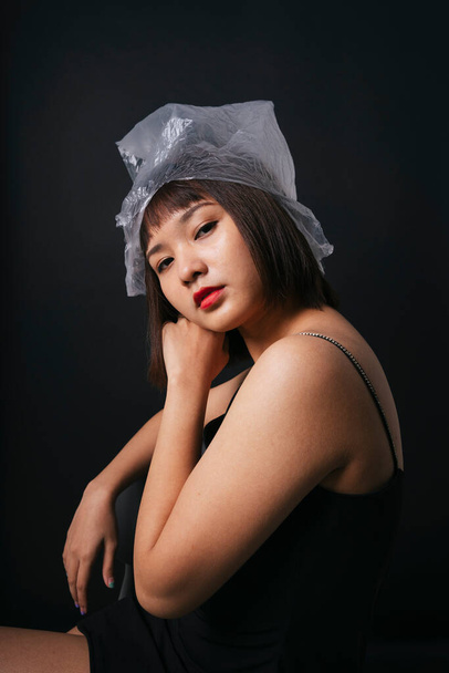 プラスチックファッションのコンセプト。美しいアジアのタイ人女性短い髪でビニール袋で隔離された黒の背景. - 写真・画像