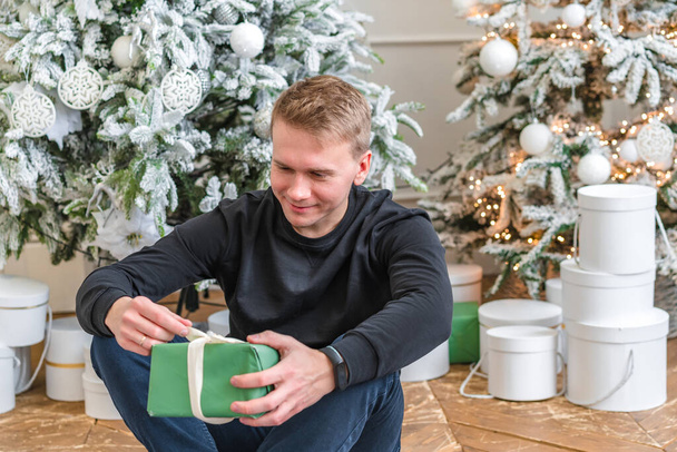 Boldog ember, szilveszteri ajándékokkal, aki a szőnyegen ül karácsonykor. Vidám fehér férfi modell karácsonykor a stúdióban. - Fotó, kép
