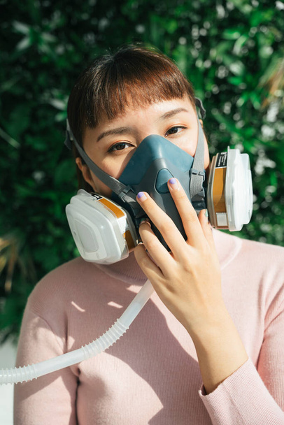 Beautiful asian thai woman in pink shirt wearing respirator gas mask for breathing during covid-19 pendemic over green background. - 写真・画像