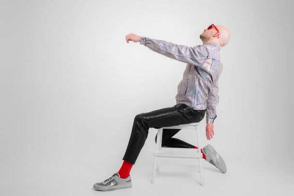 Strange caucasian man without hair in grey shirt, black leather trousers, black sunglasses, red socks and grey trainers rejoices isolated on white background - Photo, Image
