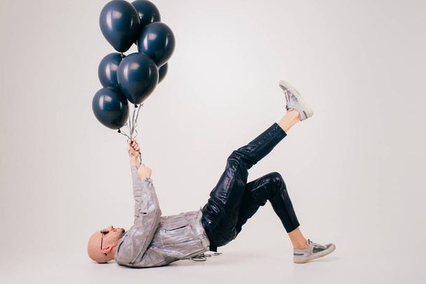 Beautiful caucasian man without hair in grey shirt, black leather trousers and grey trainers with black sunglasses lies on the floor and dream isolated on white background. - Foto, afbeelding