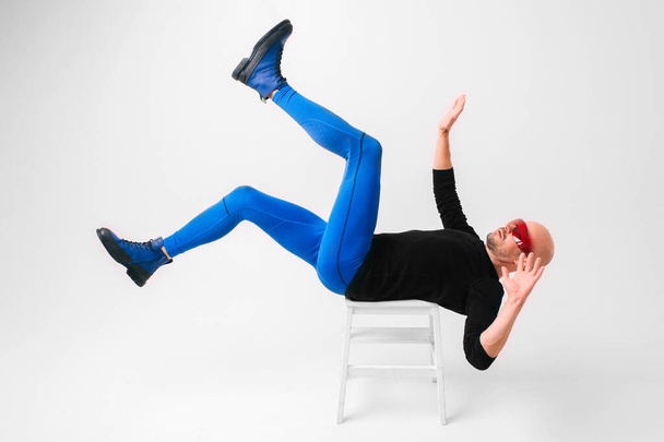 Funny and strange man in stylish clothes lying on white chair in studio in unusual pose with legs and hands apart - Foto, immagini