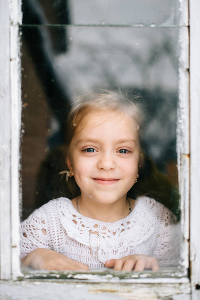 Μικρό χαρούμενο κορίτσι με χαμογελαστό πρόσωπο κοιτάζοντας κάμερα μέσα από τα παράθυρα με αποσπά αντανακλάσεις. - Φωτογραφία, εικόνα