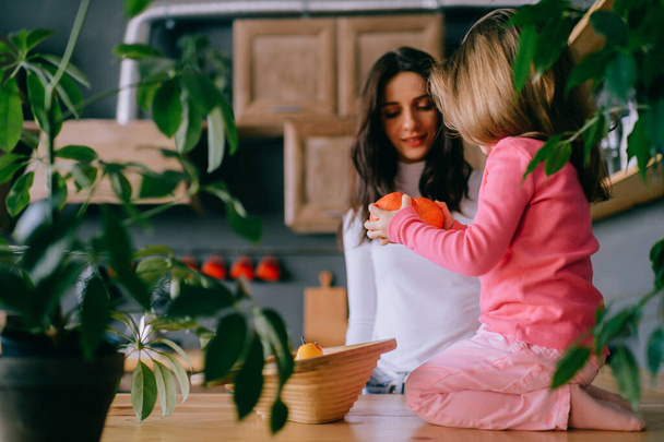 Adorable young woman playing with her little funny daughter in kitchen. Portrait of pretty mother hugging, carrying and watching her litle female child. Happy family indoor lifestyle portrait. - Photo, image