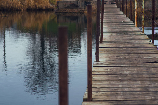 Небольшой деревянный мост через спокойную реку. Переход через плотину в виде девственной кладки. Пересекаю озеро. Мокрая, деревянная кладка осенним днем - Фото, изображение