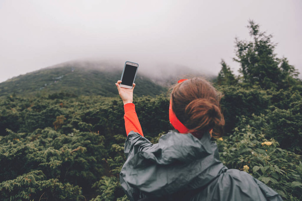 Goverla, Ukraine - July 14, 2018: Lifestyle portrait of adventure traveler girl in wet raincoat take selfie on smartphone high in foggy mountains under rain. Young female hiker take self pic on phone  - Foto, immagini