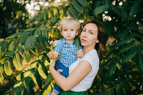 Beautiful caucasian mum with dark hair in white t-shirt and long green skirt plays in the park with her lovely son with short fair hair in white and blue shirt and blue jeans. - Foto, Imagen