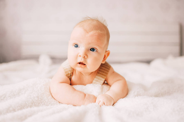 Adorable lovely newborn male baby boy with smiling emotional  happy face lifestyle indoor portrait. Funny infant child lying on stomach on bed with white wall on background.  Carefree childhood - Foto, immagini