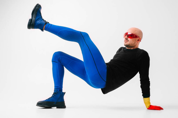 Strange man in red yellow rubber gloves in futuristic glasses and indigo color tights with boots posing over white studio background - Фото, зображення