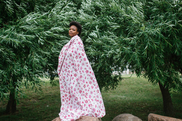 Beautiful dark skinned girl wrapped in sheet with fashion print posing at park with trees on background. - Foto, Imagen
