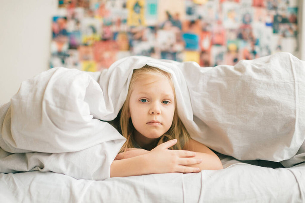 Young adorable blonde girl with funny face lying on bed under white blanket and look at camera with sad eyes. Little female child with pensive face indoor lifestyle portrait. Little  girl portrait - Фото, зображення