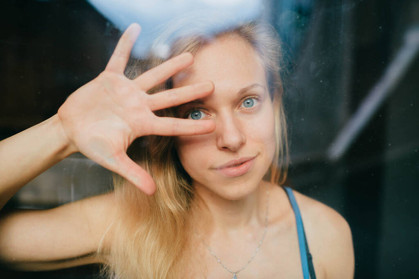 Portrait of lovely caucasian young girl with blue eyes, fair hair, pretty face behind glass and covers face with her hand and say to stop - Фото, изображение