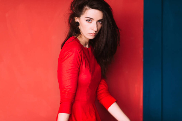 Beautiful fashion girl with dark long hair, spanish appearance and sensual face in red elegant dress posing on blue red wall in studio. Indoor lifestyle soft focus portrait of stylish brunette babe. - Photo, image
