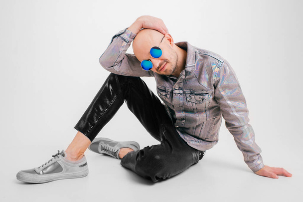 Stylish caucasian man without hair in grey shirt, black leather trousers and grey trainers with black sunglasses sits on the floor and poses for the camera isolated on white background. - Foto, Bild