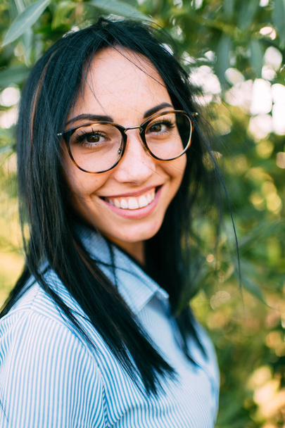 Lovely young caucasian girl with big brown eyes, long dark hair and perfect smile in short blue dress with black eyeglasses near the green tree in the garden - Fotó, kép