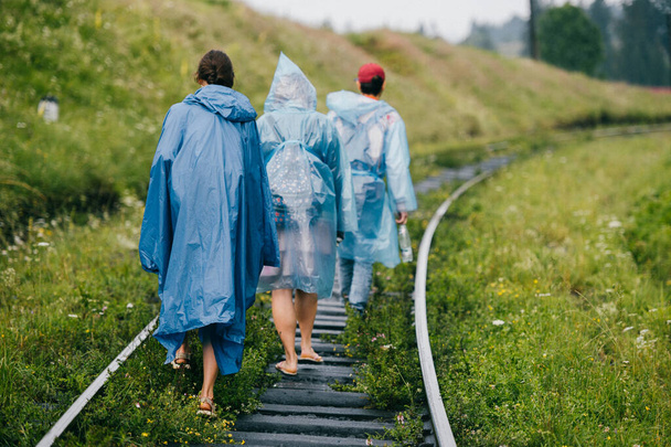 Crew of informal young travelers in raincoats walking in danger zone on railroad outdoor at nature. Company of friends rest on vacation. People go on railway sleepers. Cloudy muggy weather - Photo, Image